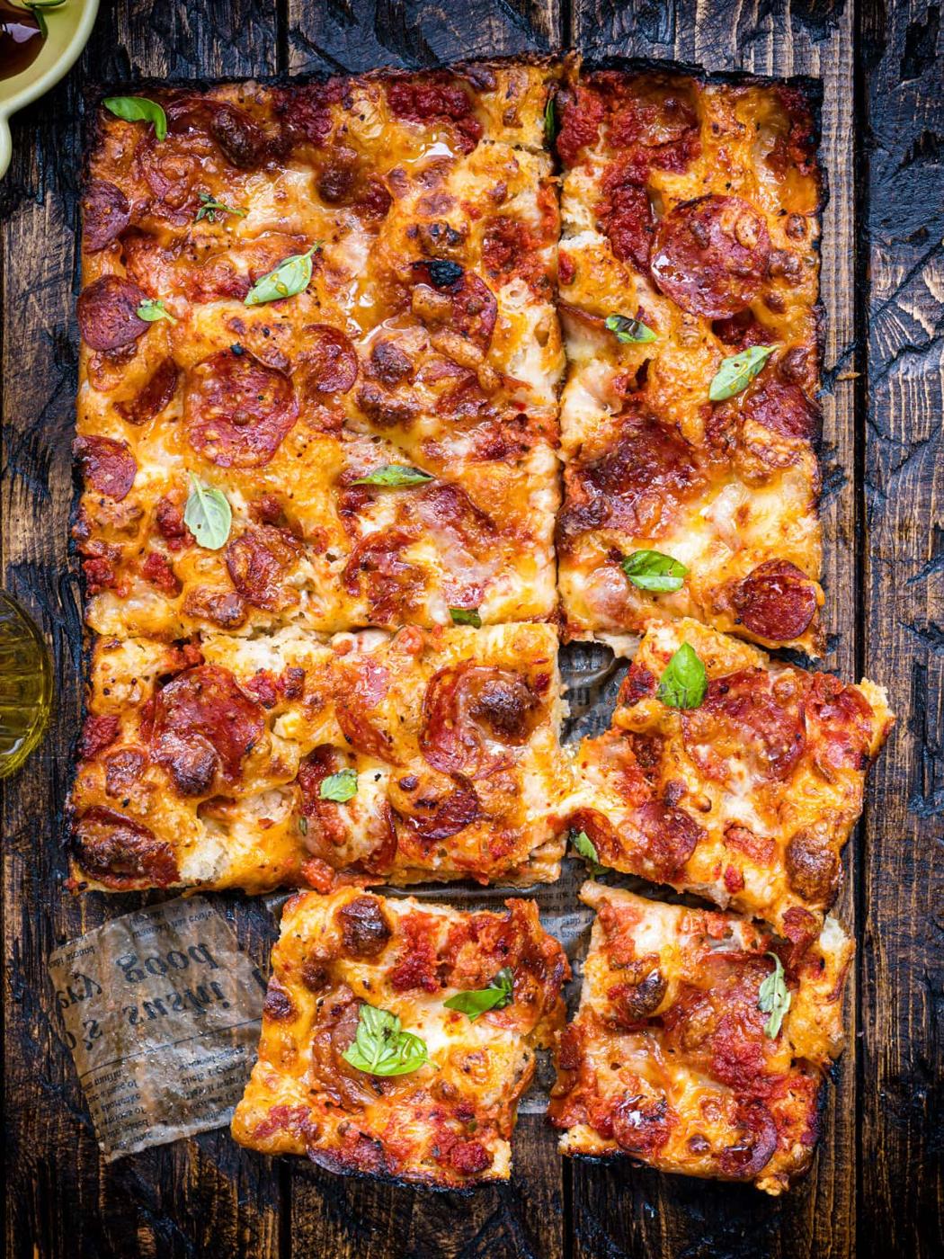 Rectangular pepperoni focaccia pizza with melted cheese and tomato sauce on a rustic wooden surface, cut into six square slices, garnished with fresh basil leaves.