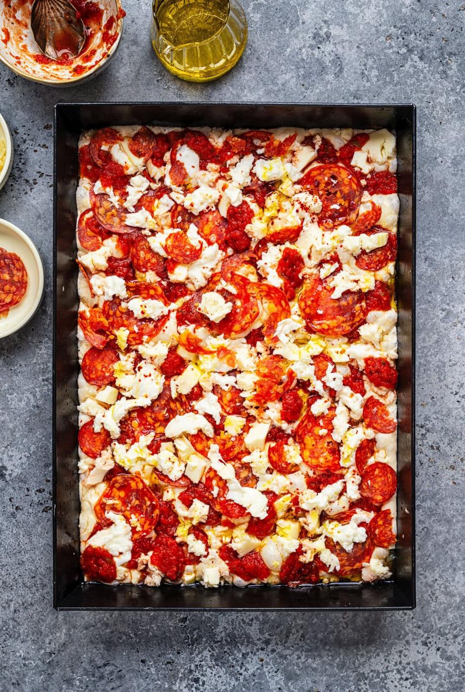 A rectangular pan filled with focaccia pizza dough topped with tomato sauce, mozzarella cheese, and slices of pepperoni, ready to be baked. A bottle of olive oil and small bowls with ingredients are nearby.