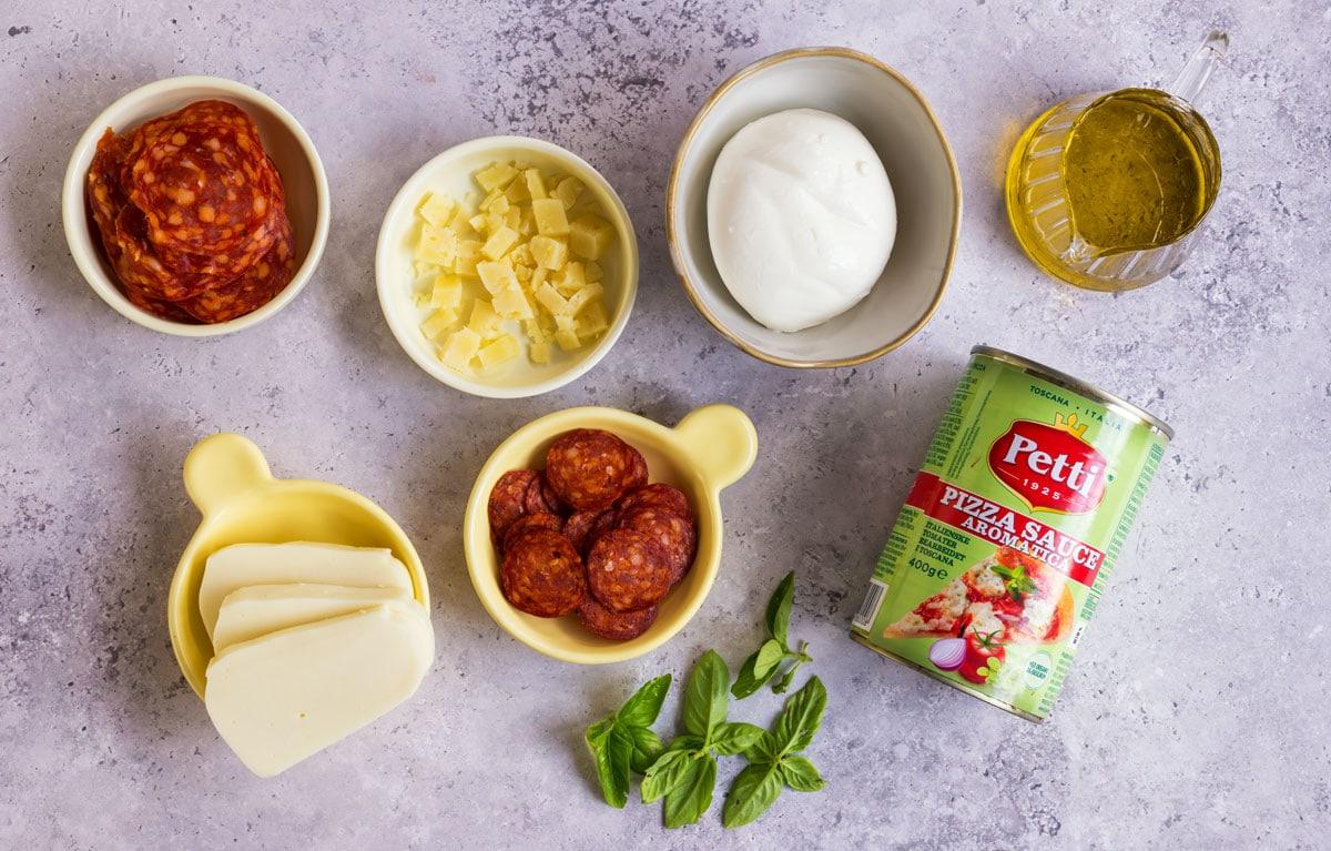 Assorted pizza ingredients on a light surface: sliced cheese, cubed cheese, mozzarella ball, sliced pepperoni, sliced chorizo, fresh basil, a can of pizza sauce, and a glass container of olive oil.
