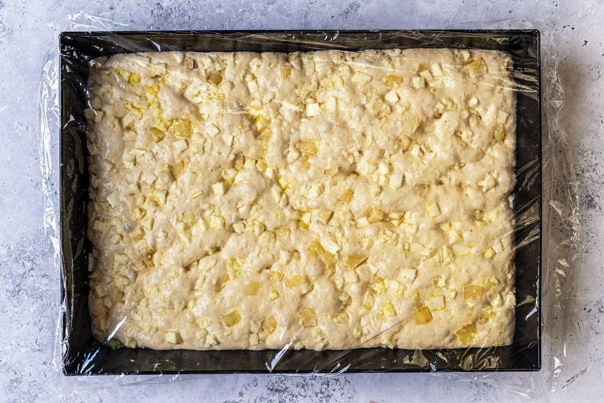 A rectangular baking tray filled with unbaked dough, topped with small butter pieces and loosely covered with a sheet of plastic wrap, sits on a light gray textured surface.