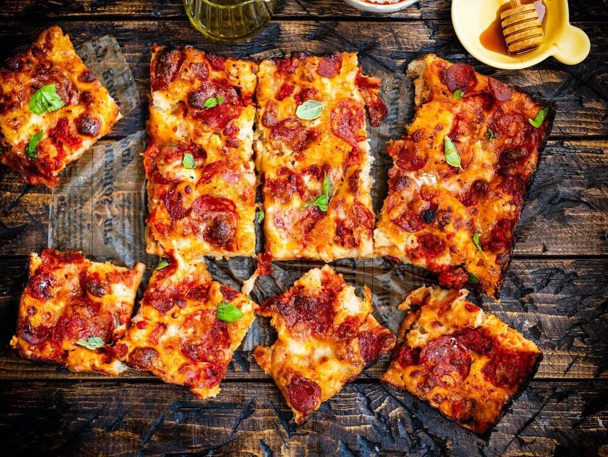Rectangular slices of pepperoni pizza with melted cheese and fresh basil are arranged on rustic parchment paper over a wooden table. A small dish of honey and a glass of olive oil sit nearby.