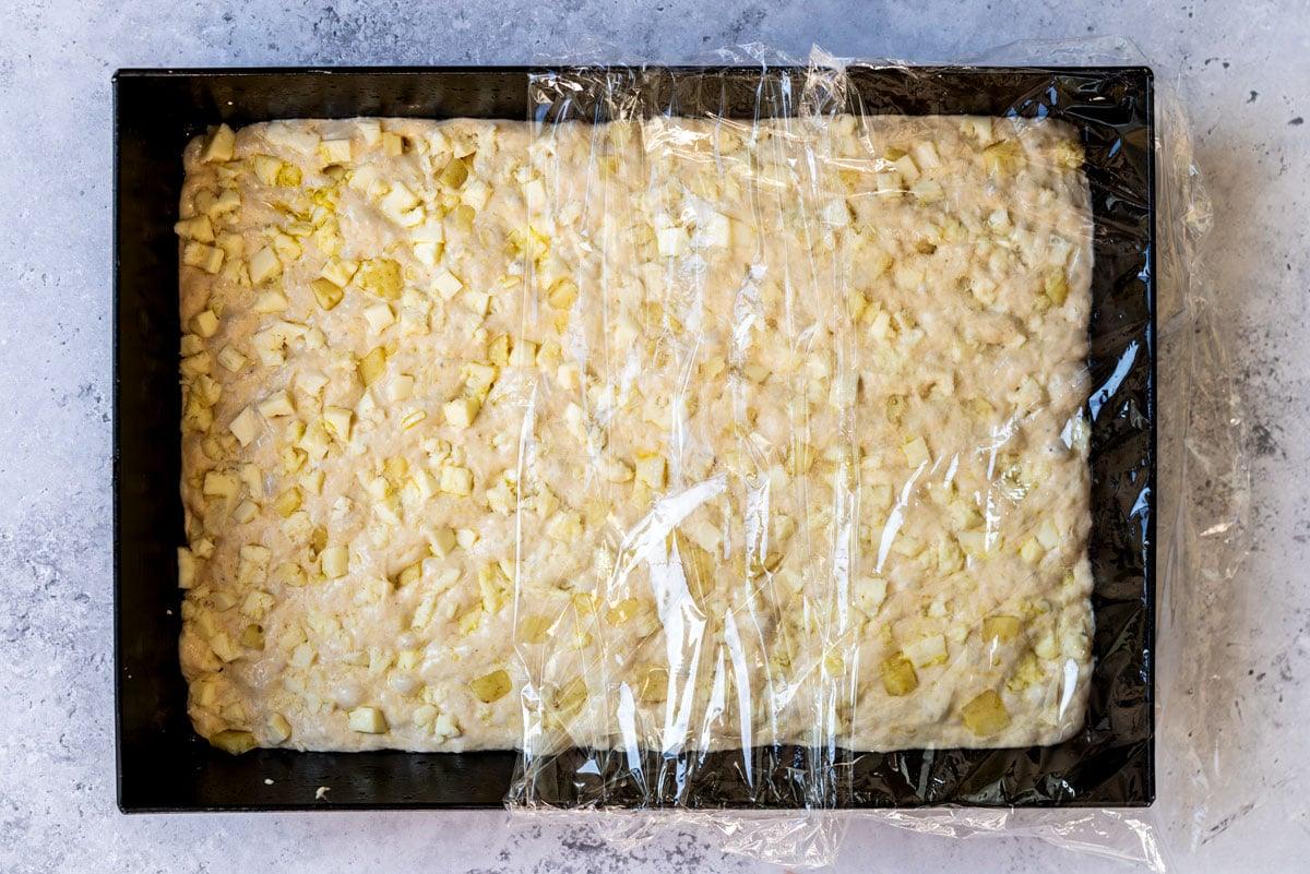 A rectangular baking pan filled with dough mixed with apple chunks, partially covered with clear plastic wrap, sits on a light gray surface.