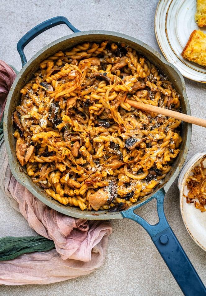 A skillet filled with French onion pasta with mushrooms, chicken sits on a light countertop, with a wooden spoon resting inside. Plates and colorful cloth napkins are nearby.