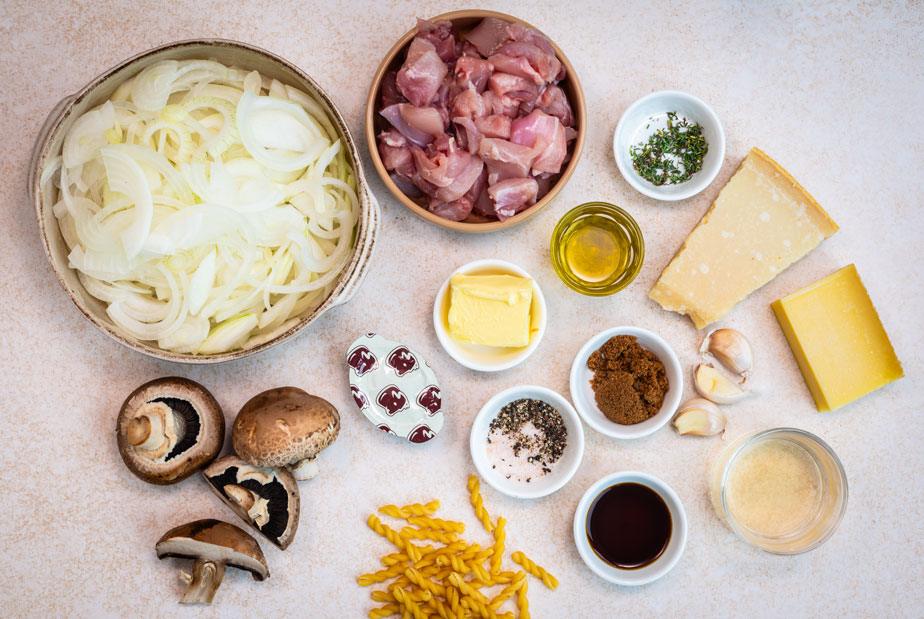 A variety of ingredients on a light surface, including sliced onions, cubed raw chicken, cheese wedges, butter, olive oil, garlic cloves, spices, mushrooms, dry pasta, and small bowls with liquids and seasonings.