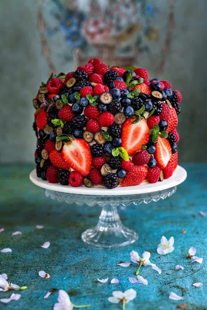 A fresh berry cake is covered with strawberries, blueberries, raspberries, and blackberries, then decorated with small pink flowers. It rests on a round white plate against a blue background.
