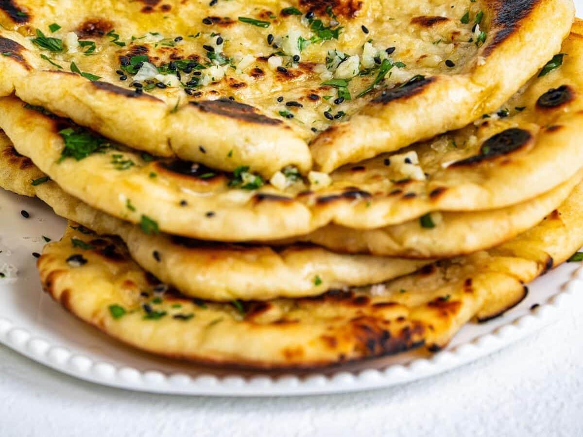 A stack of golden-brown naan bread topped with chopped herbs and garlic, served on a white plate against a light background.