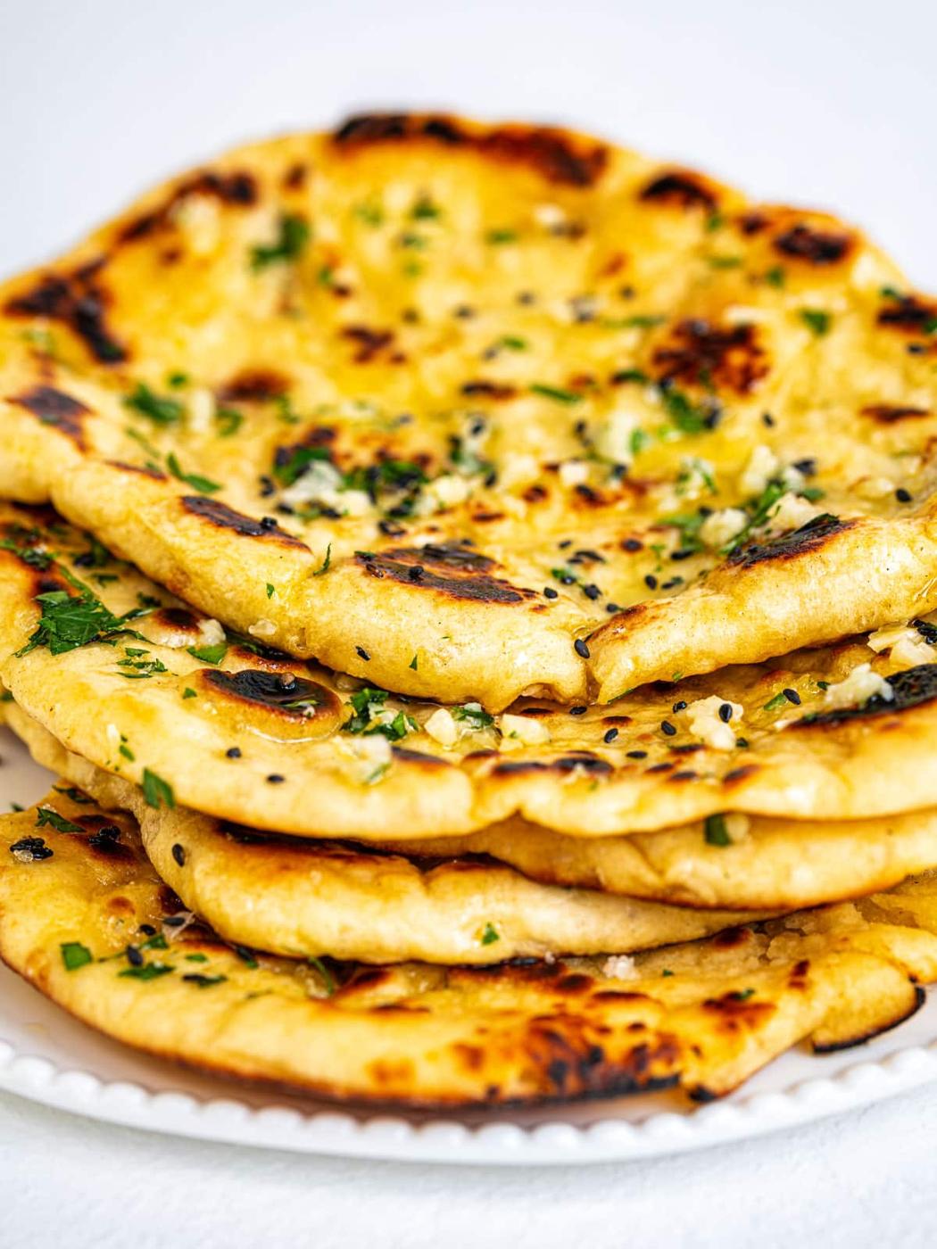 A stack of fresh Indian garlic naan bread topped with chopped herbs and garli butter, displayed on a white plate.