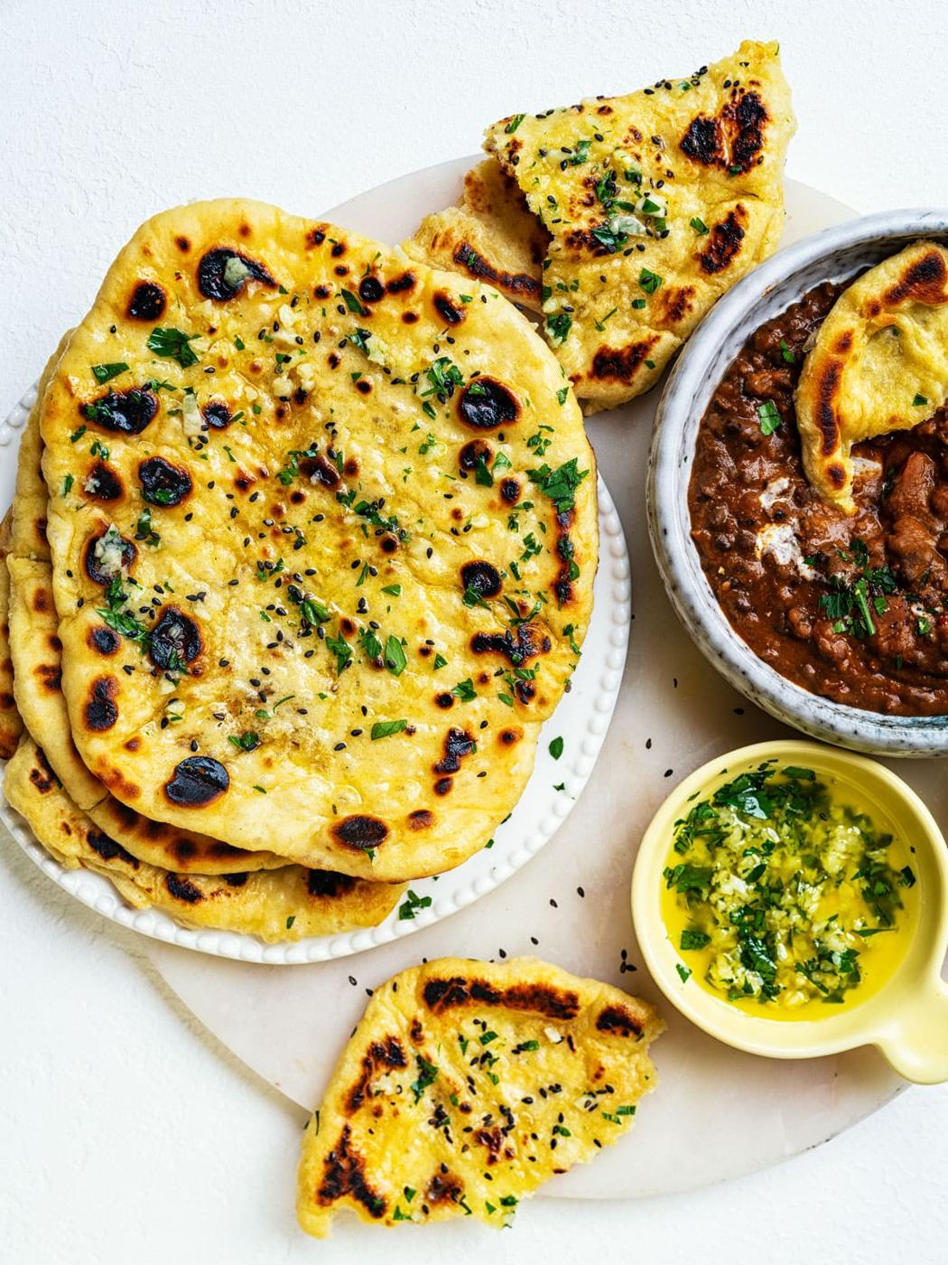 A plate of homemade garlic naan bread garnished with herbs is served alongside a bowl of dal and a small dish of green herb sauce, all presented on a white surface.