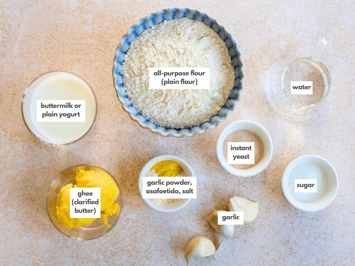 Various naan bread ingredients are arranged on a countertop, including labeled bowls of all-purpose flour, buttermilk or yogurt, ghee, garlic powder with spices, instant yeast, water, sugar, and whole garlic cloves.