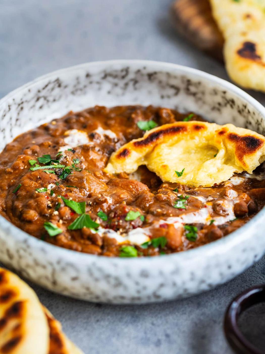 A bowl of creamy lentil daal garnished with herbs, with a piece of naan bread partially dipped in the curry. The background shows more naan and a small dish on a gray surface.