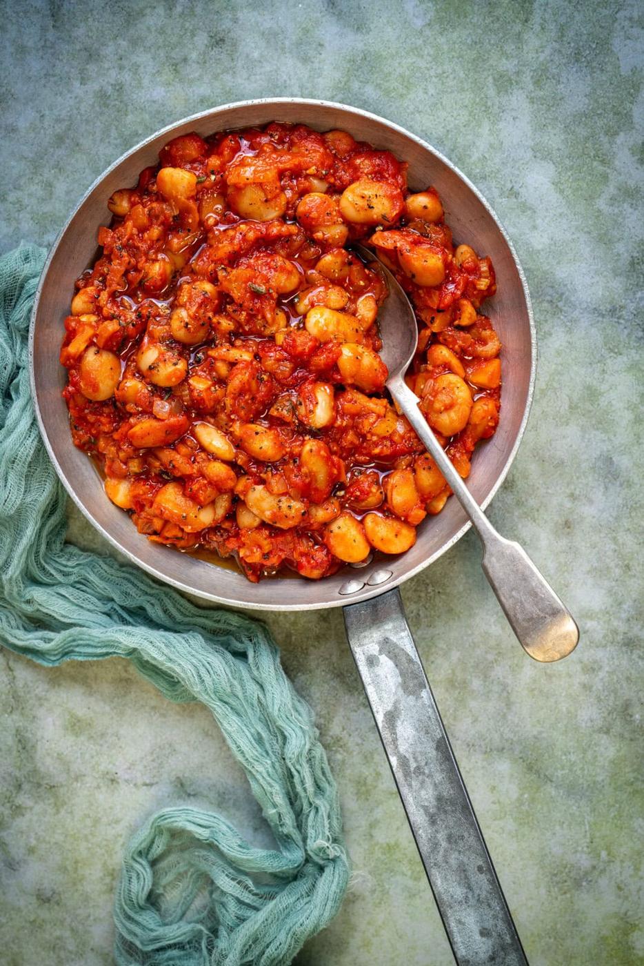 Overhead view of a pan filled with Greek giant beans in a rich tomato sauce, garnished with herbs. A spoon rests inside the pan, and a textured green cloth lies beside it on a grayish surface.