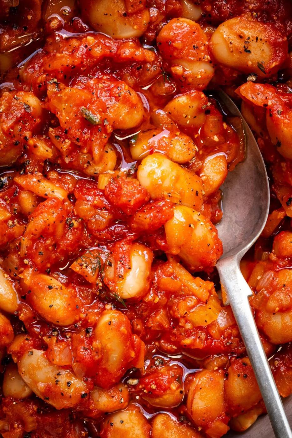Close-up of a spoon resting in a dish of stewed gigantes beans in a chunky tomato sauce, seasoned with herbs and black pepper.