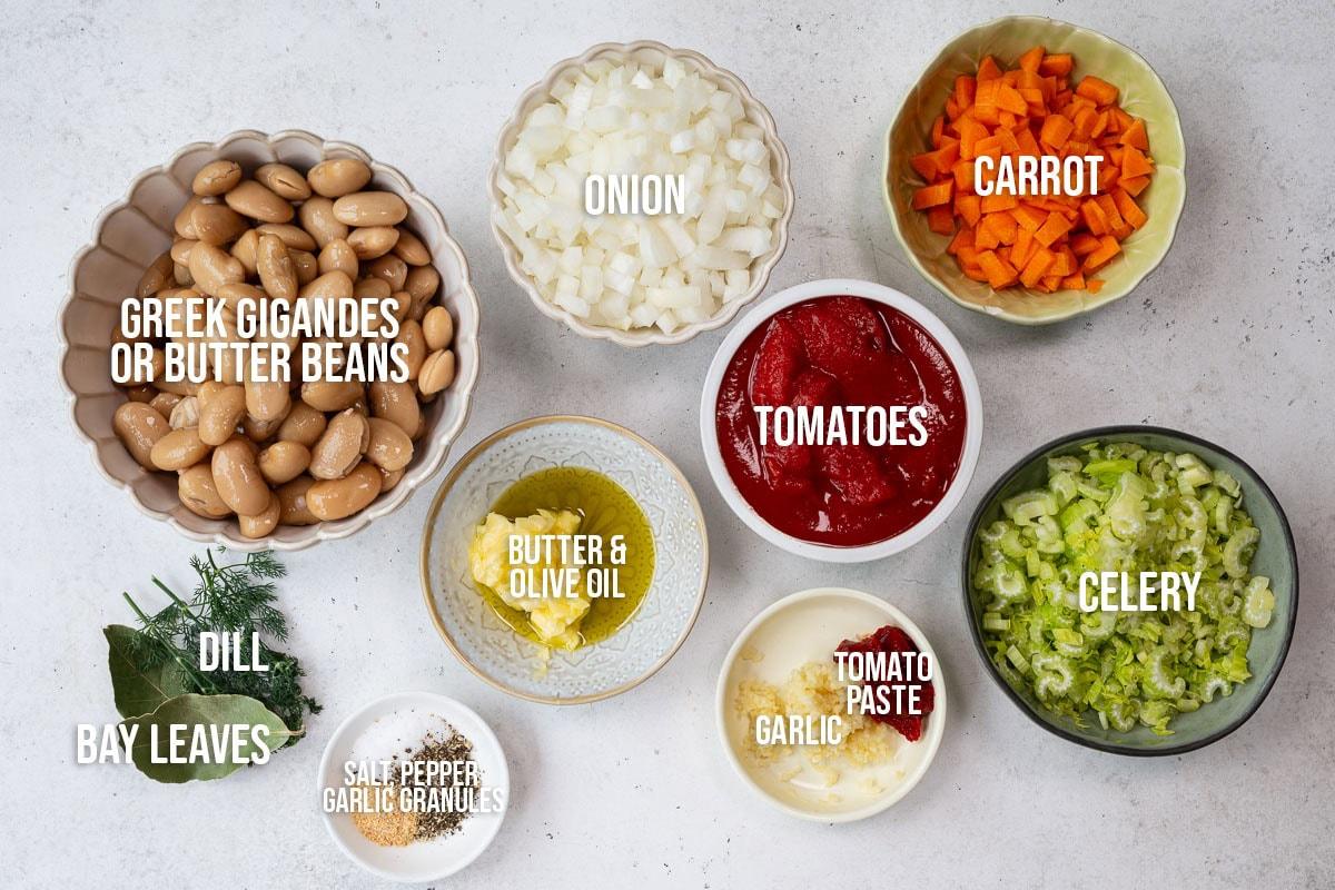 A top-down view of bowls containing chopped onion, carrot, celery, tomatoes, Greek gigantes or butter beans, dill, bay leaves, butter and olive oil, tomato paste, garlic, and a dish of salt, pepper, and garlic granules.