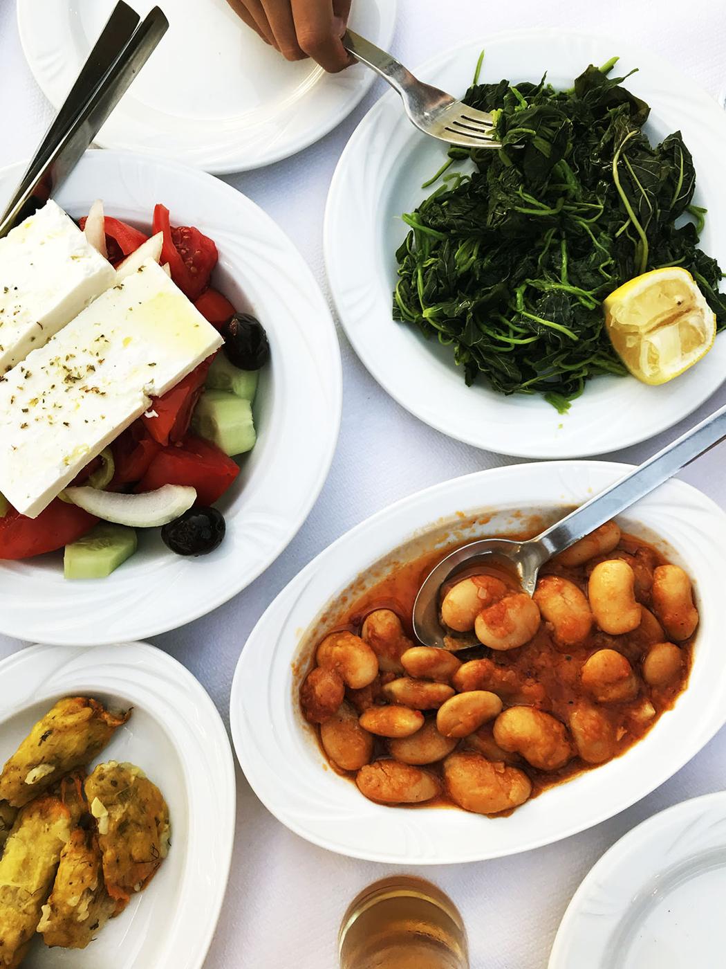 A table with Greek dishes: a salad with feta, tomatoes, cucumbers, and olives; a plate of cooked greens with a lemon wedge; baked giant beans in tomato sauce; and fritters on small plates. A hand holds a fork.