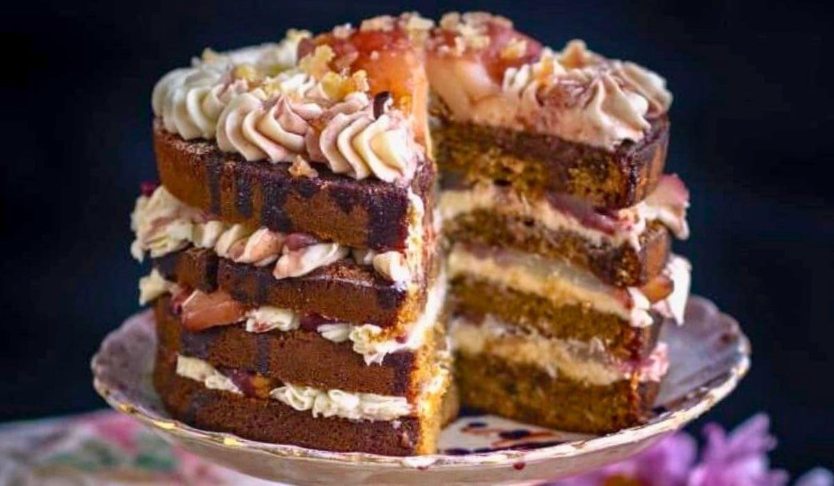 A four-layer Pear Cake with cream frosting and fruit topping sits on a cake stand. A large slice has been removed, revealing layers of cake, cream, and fruit filling inside—perfect as a stunning Thanksgiving Cake centerpiece.