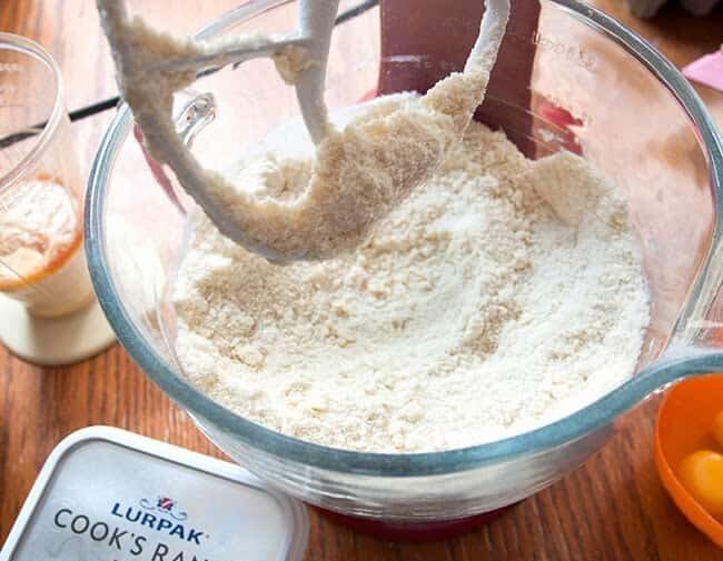 A glass mixing bowl filled with flour and a stand mixer attachment covered in flour. Nearby are a tub of butter, a glass of liquid, and an orange bowl with egg yolks.