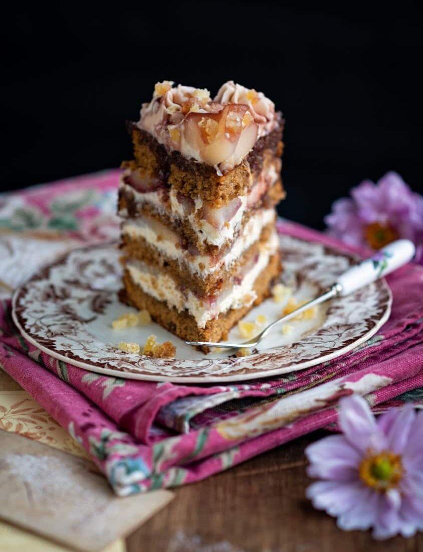 A tall slice of layered Pear cake with frosting and fruit topping sits on a decorative plate, atop stacked pink napkins. This elegant dessert is paired with a floral-handled fork and purple flowers on the wooden table—perfect for Thanksgiving.