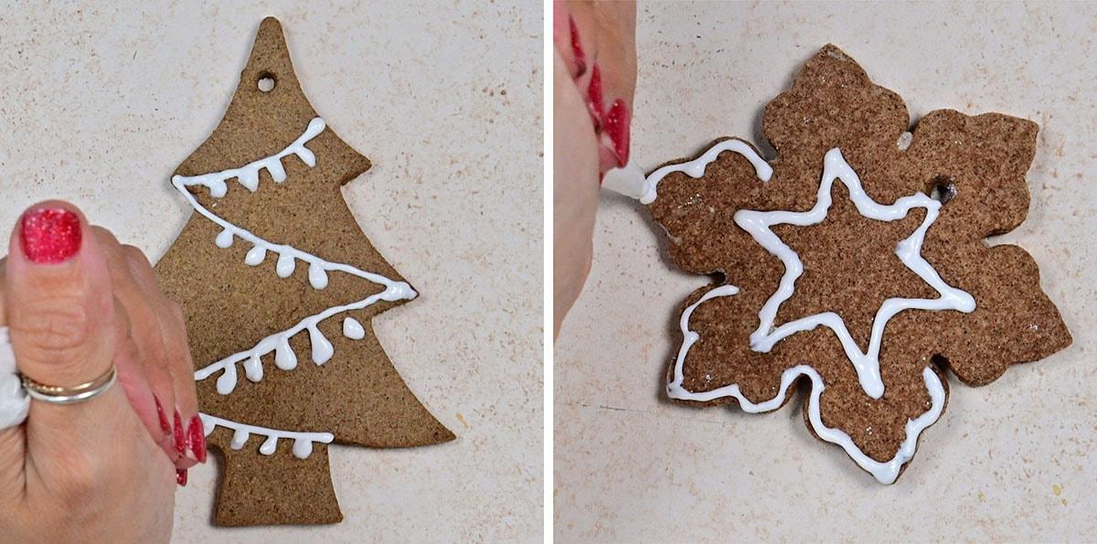 Two hands decorate Christmas gingerbread ornaments with white icing; one is a Christmas tree with garland, the other is a snowflake with star and outline patterns. Both cookies are on a light countertop.