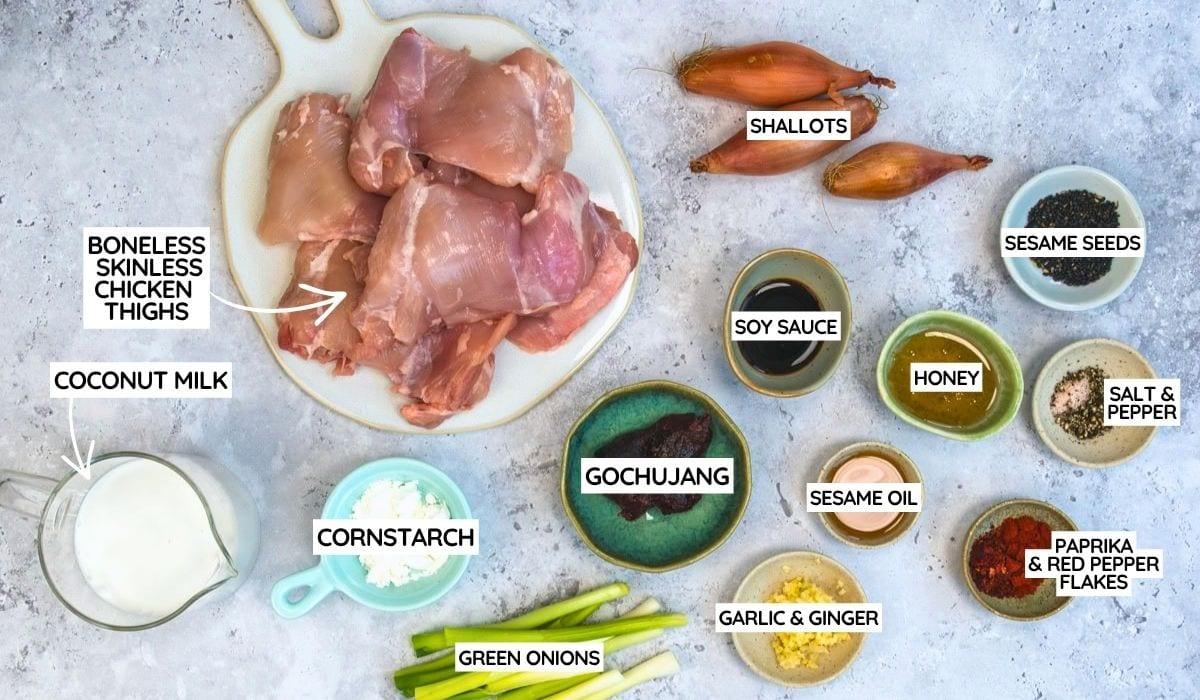 A flat lay of labeled ingredients for a chicken dish, including boneless skinless chicken thighs, coconut milk, shallots, soy sauce, honey, sesame oil, gochujang, cornstarch, green onions, garlic & ginger, sesame seeds, salt & pepper, and paprika & red pepper flakes.
