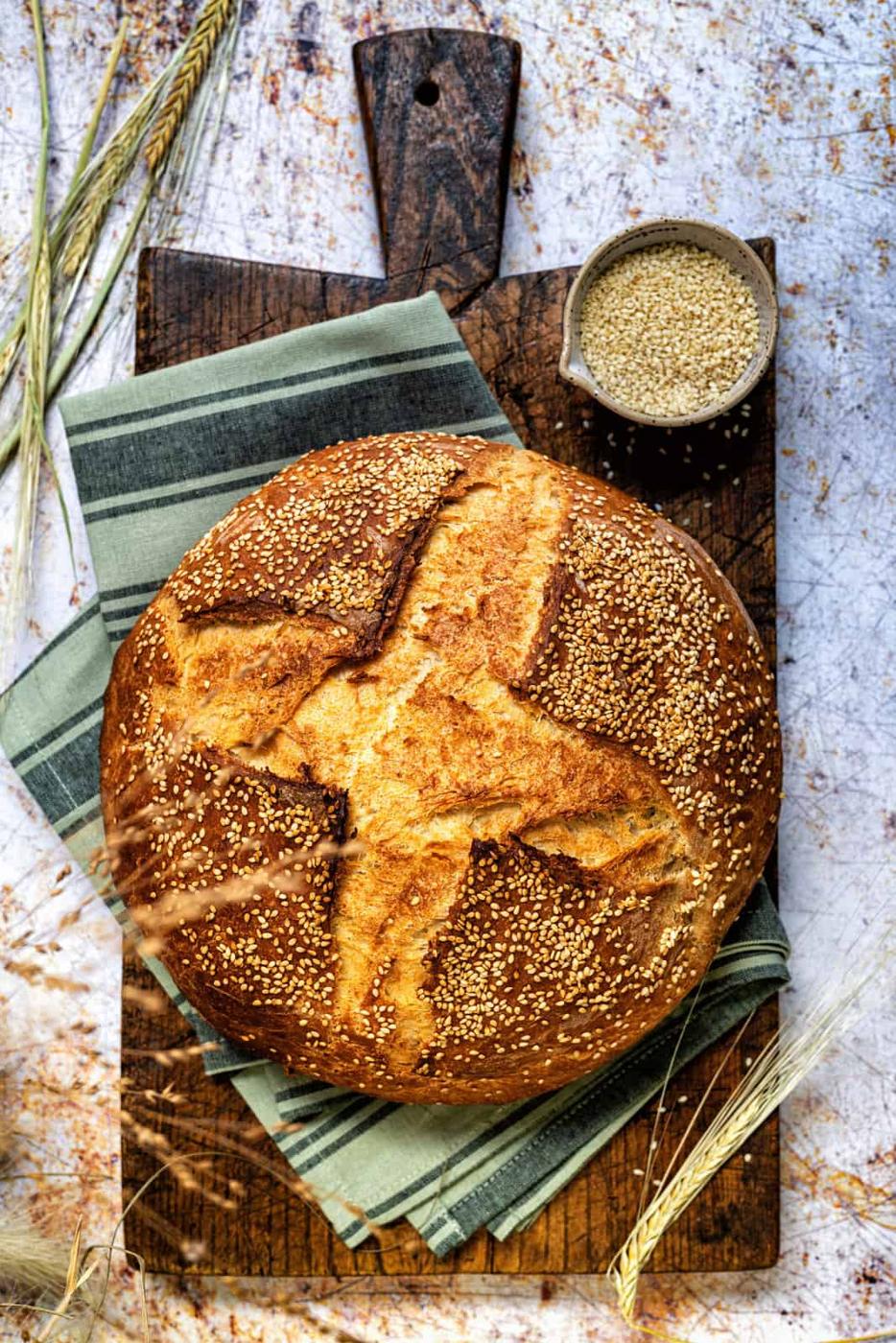 Greek Country Style Village bread topped with sesame seeds on a wooden board, small bowl of sesame seeds on the side