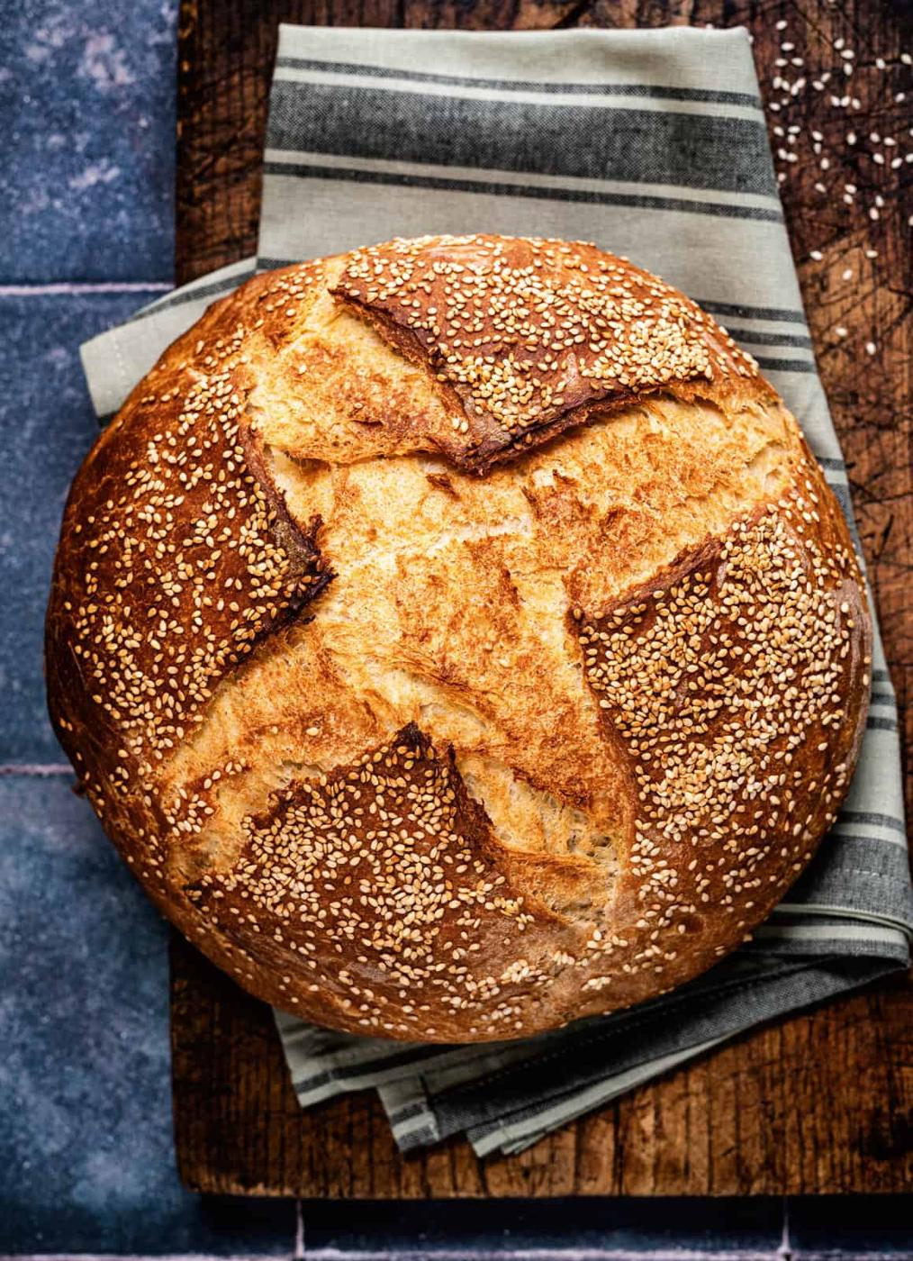 A round loaf of Greek village bread with a crusty top and sesame seeds sits on a striped cloth on a wooden surface, its golden-brown aroma invoking memories of traditional recipes.
