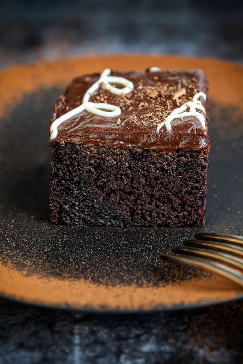 A slice of rich chocolate cake (sokolatopita) with smooth chocolate frosting and white icing details on top. The cake is placed on a plate dusted with cocoa powder. A fork is positioned in the foreground.