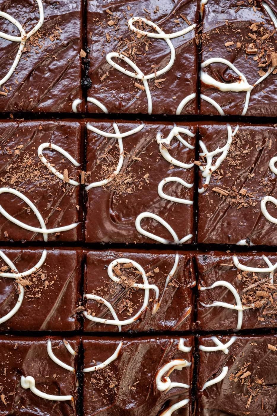 Close-up of a Greek chocolate cake topped with glossy chocolate icing and white chocolate drizzle. The cake is divided into evenly cut squares. Shavings of chocolate are scattered on top, enhancing the rich, textured appearance.