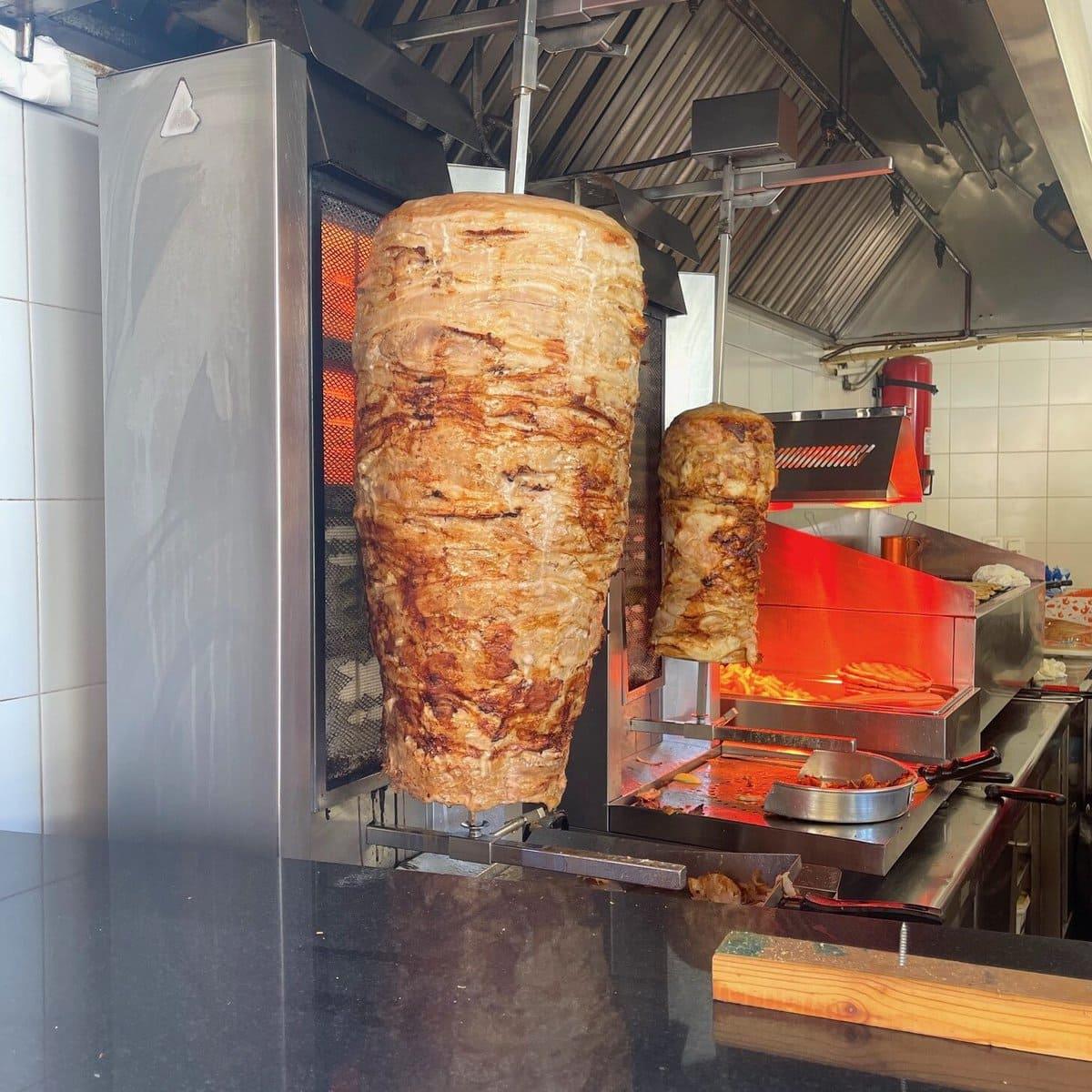 A kitchen scene showcases two vertical rotisserie spits with seasoned meat, as a succulent lamb gyro rotates on the larger spit in the foreground. The smaller one spins in the background, both sizzling and cooking perfectly near an open flame.