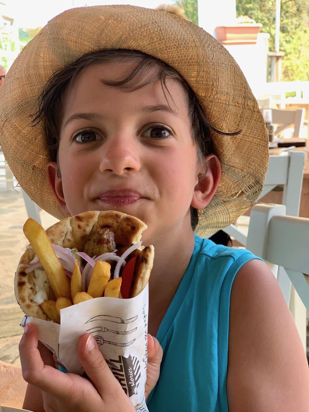 A child with short dark hair and a straw hat is holding a pita wrap filled with fries, onions, and other ingredients for a classic lamb gyro. They are wearing a blue shirt and sitting at an outdoor table, smiling at the camera.