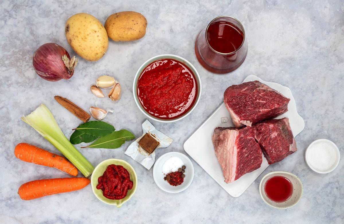 Raw ingredients for a stew, including potatoes, red onion, garlic, carrots, celery, beef chunks, tomato paste, tomato sauce, red wine, seasonings, and bay leaves arranged on a light surface.