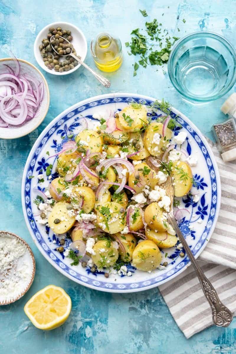 A bowl of Greek potato salad with red onions, feta cheese, herbs, and capers on a blue-patterned plate, surrounded by lemon, salt, pepper, oil, and a striped serviette on a blue table.