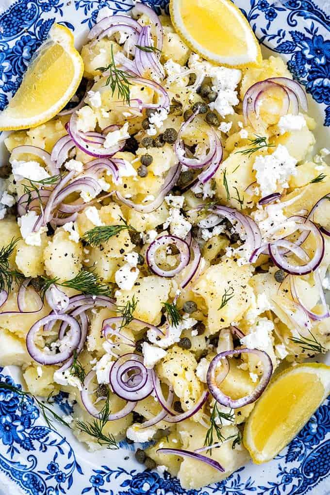 A blue and white plate with a potato salad topped with sliced red onions, capers, dill, crumbled feta cheese, black pepper, and lemon wedges.