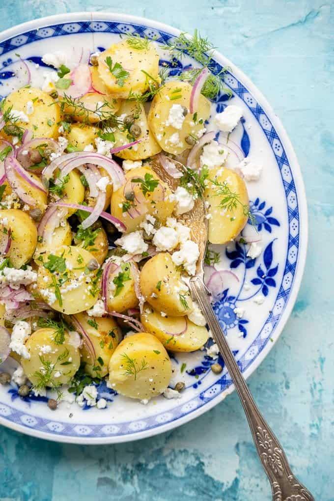 A blue and white plate filled with Greek Potato Salad made of sliced baby potatoes, red onion, crumbled feta cheese, capers, and fresh herbs, with a silver serving spoon on the side.