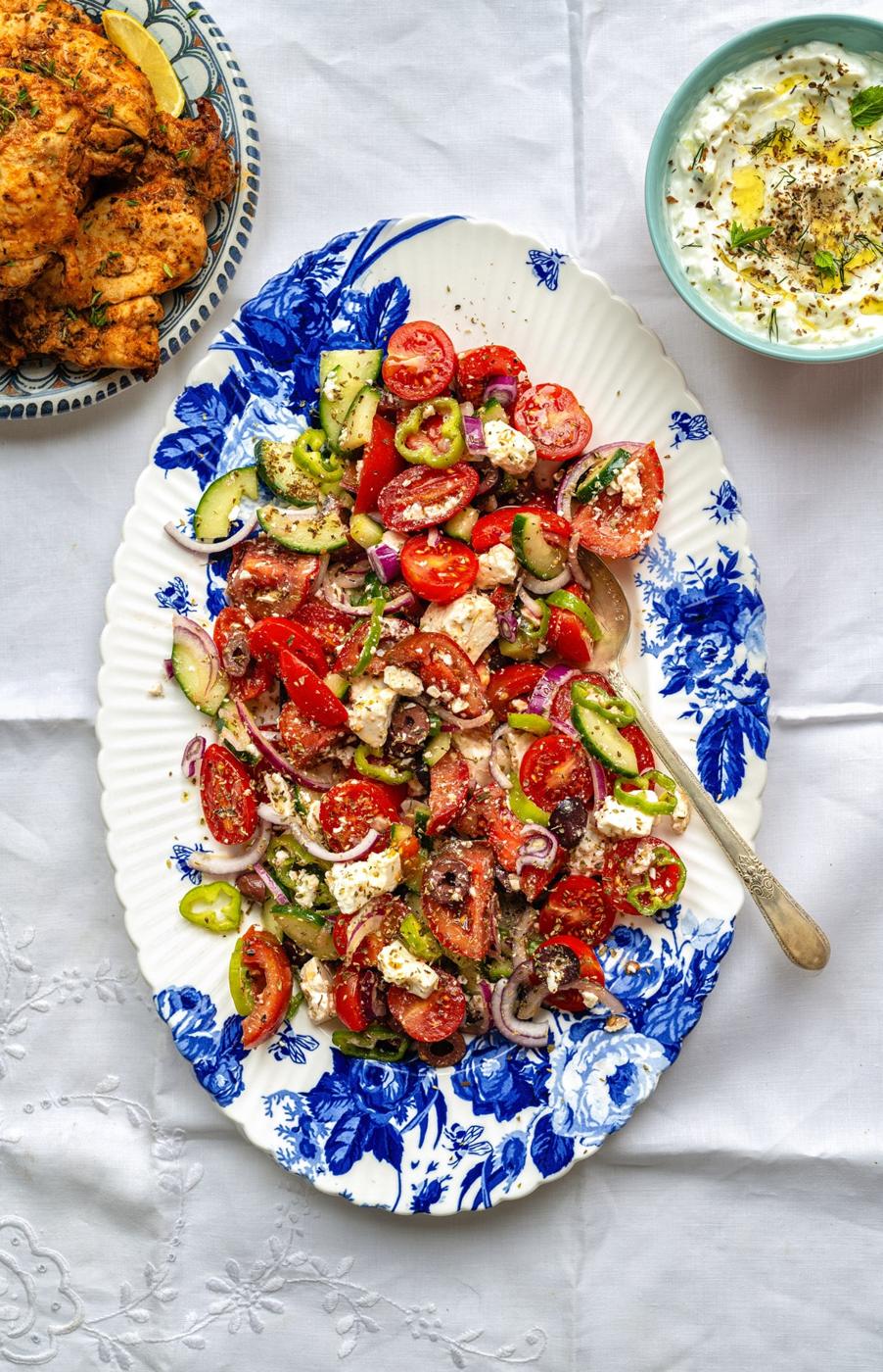 Greek salad on a blue and white floral platter with tzatziki sauce and grilled chicken on the side