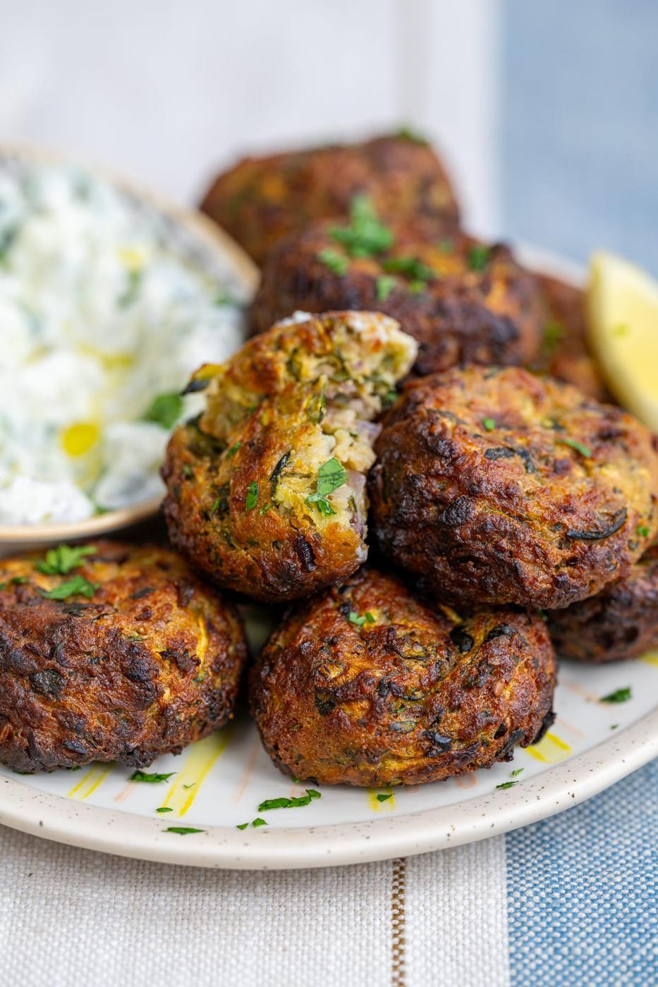 A plate of golden-brown courgette fritters, known as kolokithokeftedes, garnished with chopped herbs and served with creamy dipping sauce and a lemon wedge. One fritter is broken open, revealing a moist, green and white interior.