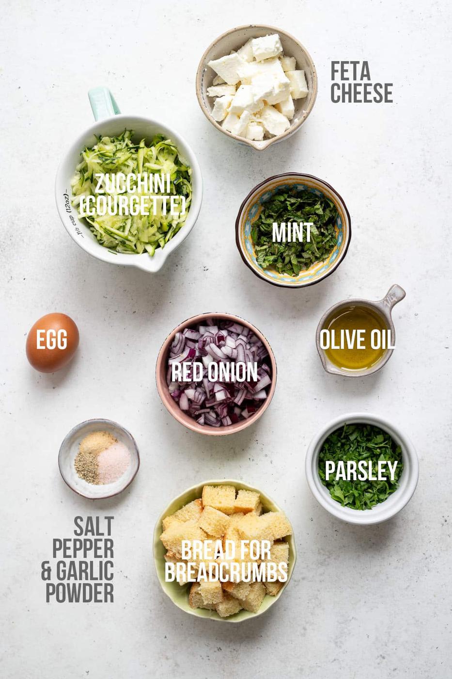 Top-down view of ingredients in separate bowls labeled: feta cheese, zucchini (courgette) for kolokithokeftedes, mint, olive oil, parsley, red onion, bread for breadcrumbs, egg, and a mix of salt, pepper, and garlic powder.