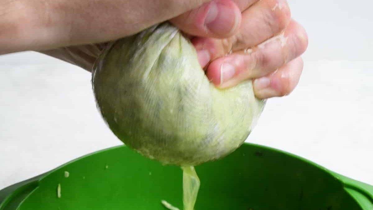 A hand squeezing shredded zucchini in a cloth, with liquid dripping into a green bowl—perfect prep for courgette fritters or Greek Zucchini Balls, also known as kolokithokeftedes.
