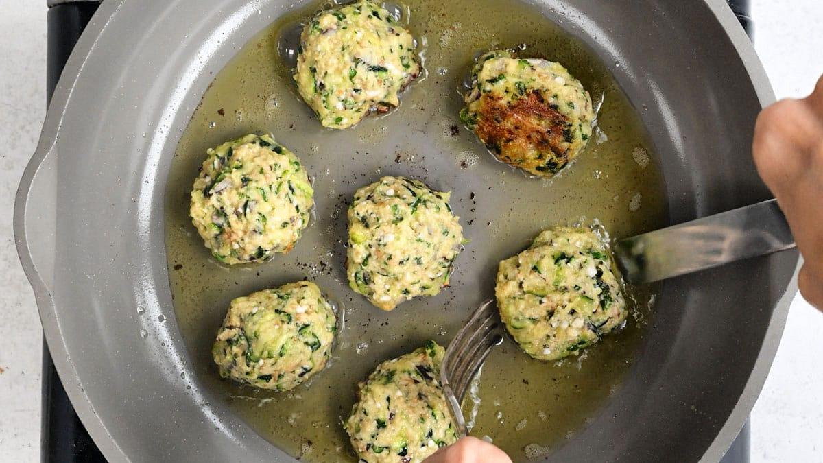 Seven kolokithokeftedes, or Greek Zucchini Balls, are being cooked in a frying pan with oil. One fritter is being flipped with a fork and spoon, revealing a golden-brown underside while the rest remain uncooked on top.