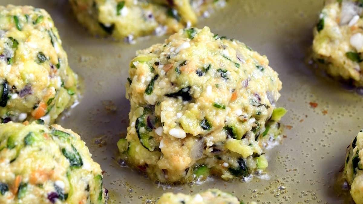 Close-up of several uncooked Greek Zucchini Balls, also known as kolokithokeftedes, made with chopped greens, carrots, and cheese, sitting on a baking sheet with a light coating of oil.