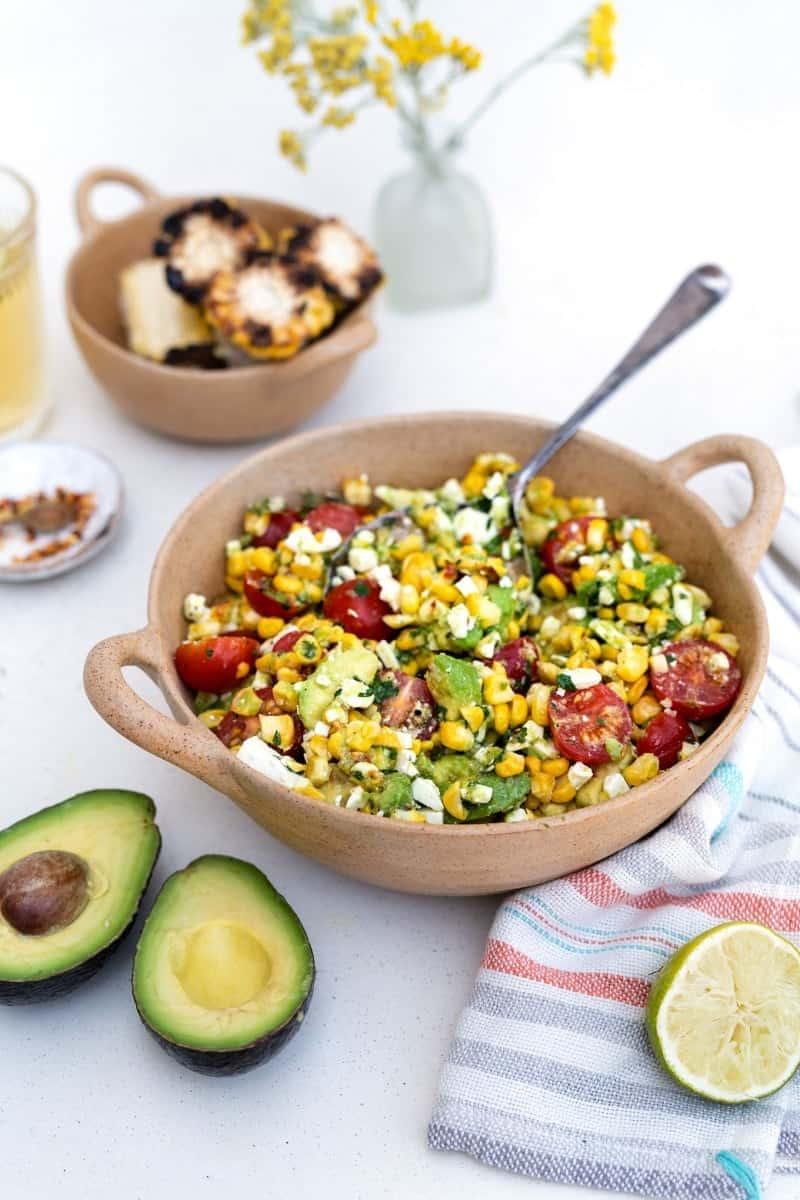 A bowl of grilled corn avocado salad with cherry tomatoes, feta, and herbs sits on a white table. Nearby are a halved avocado, lime, grilled corn slices, a drink, and a vase with yellow flowers.