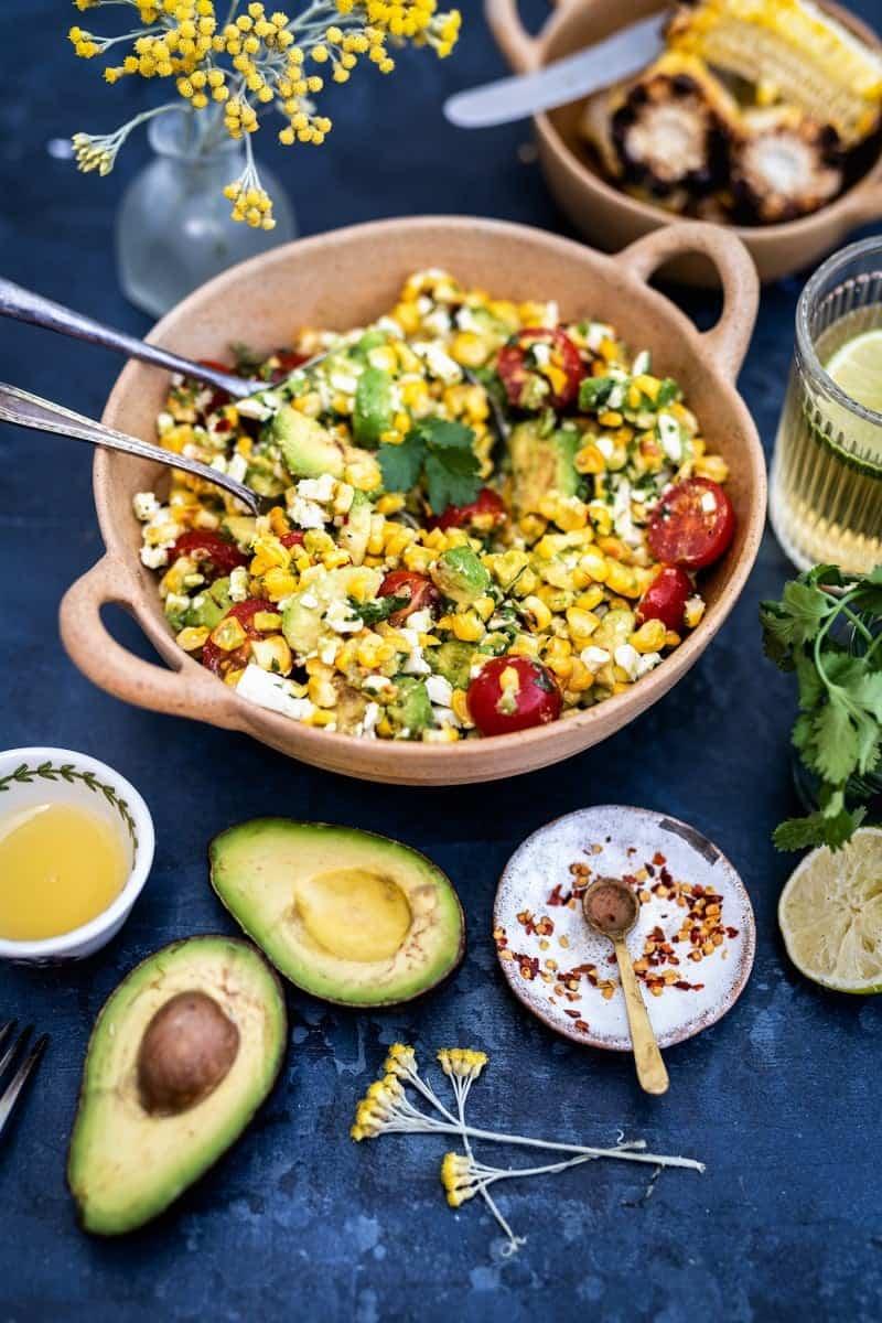 A bowl of grilled corn avocado salad with tomatoes, feta, and herbs sits on a table with cut avocado, corn, lime, spices, a glass of lemonade, and yellow flowers nearby.