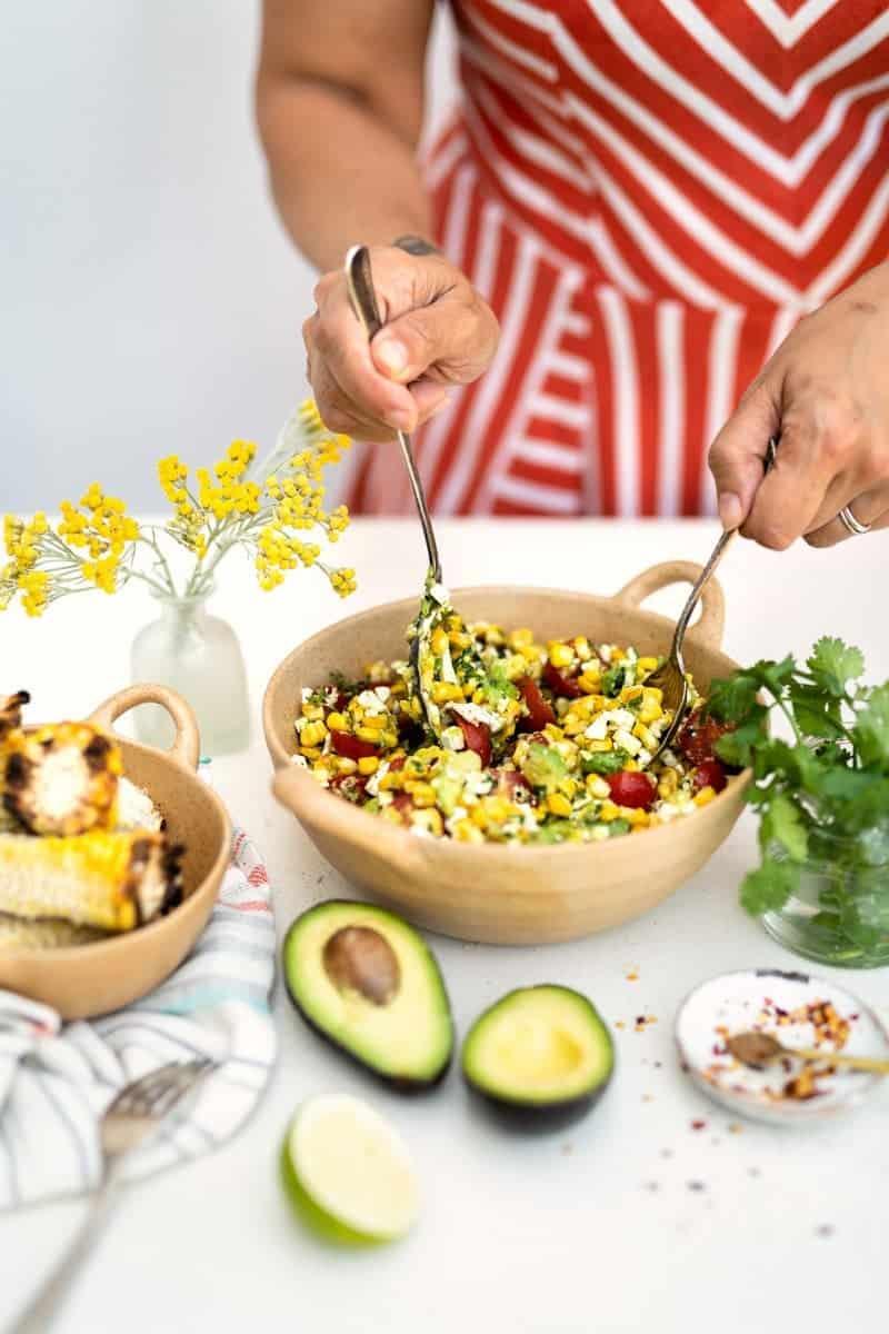 A person in a red and white striped dress mixes a vibrant grilled corn avocado salad with tomatoes and herbs in a bowl. Nearby are fresh avocados, grilled corn, a lime half, herbs, and a vase with yellow flowers.