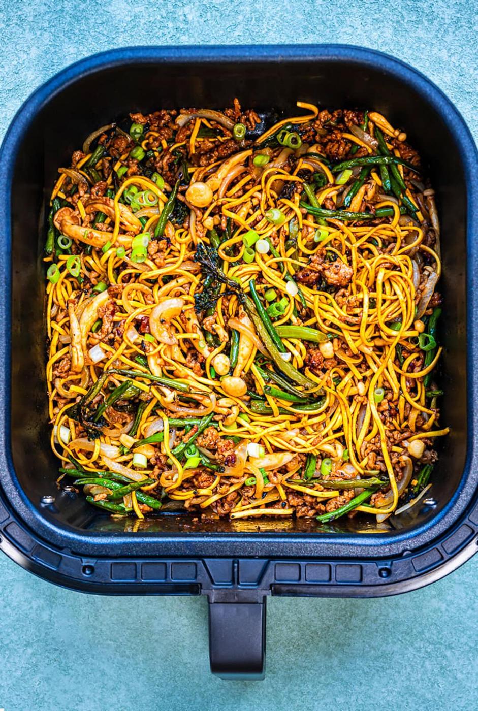 Stir-fried noodles with ground meat, green onions, green beans, and onions in an air fryer basket, viewed from above on a teal surface.