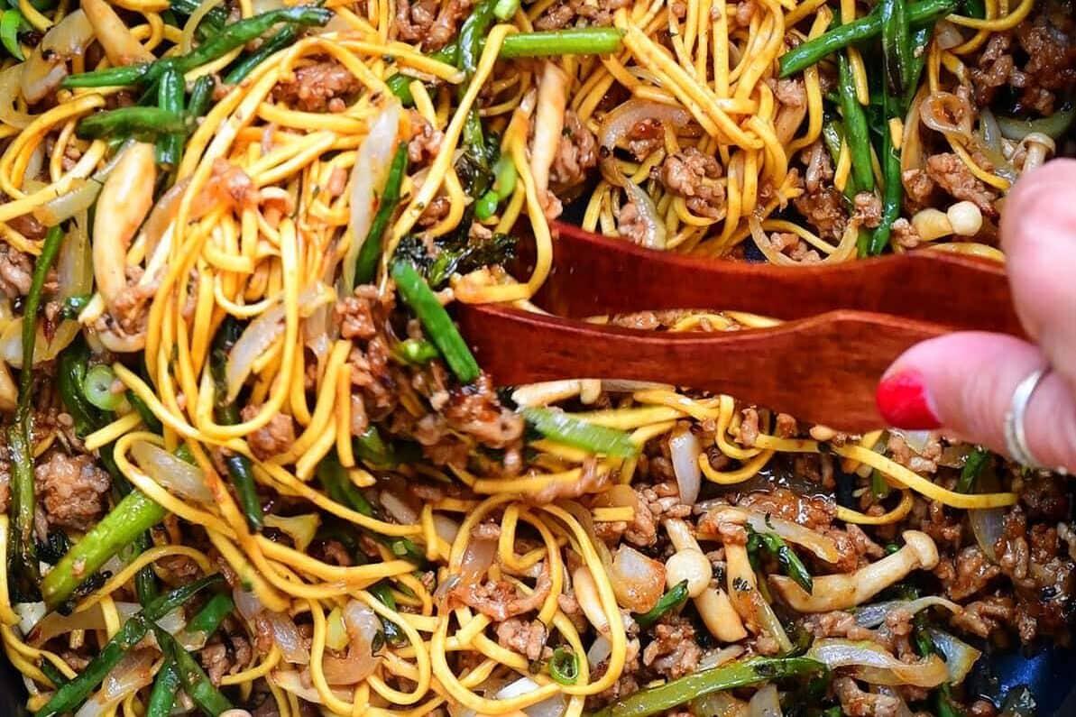 A hand uses wooden tongs to mix stir-fried noodles with ground meat, sliced onions, and green vegetables inside an air fryer basket.