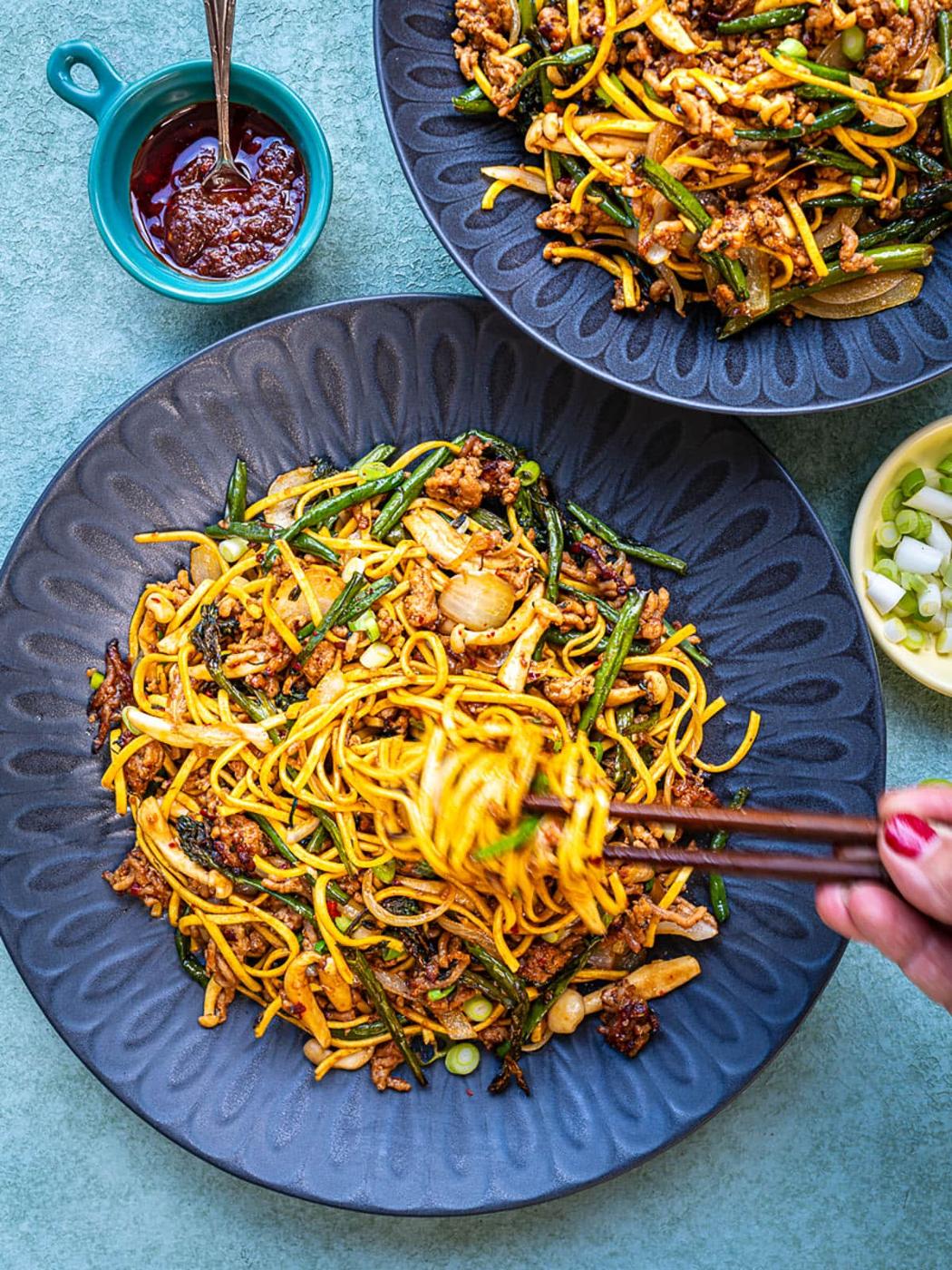 A hand holding chopsticks lifts noodles from a bowl of stir-fried noodles with vegetables and meat; another bowl, a dish of sauce, and a small bowl of chopped green onions are nearby on a blue-green surface.