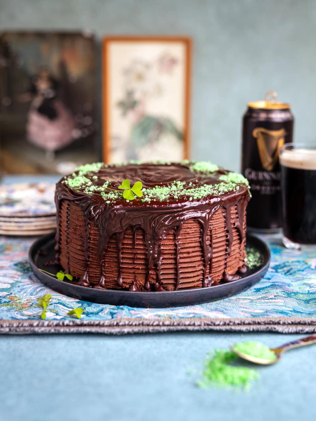 A rich Guinness chocolate cake with dripping chocolate glaze, decorated with green sprinkles and clover leaves, sits on a black plate. A can and glass of Guinness are in the background, along with framed art and stacked plates.