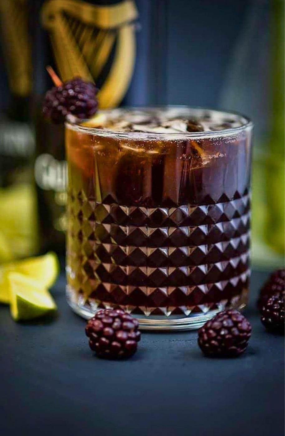 A Guinness Black Magic cocktail over ice in a diamond-cut rocks glass, garnished with a blackberry on a cocktail stick, with a Guinness bottle, lime wedges and blackberries around it.