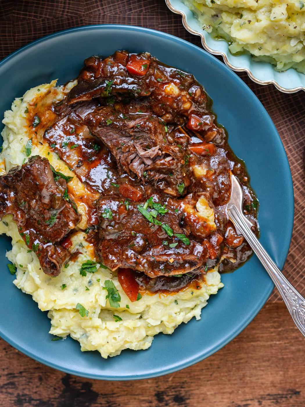 A blue plate with creamy mashed potatoes topped with slow-cooked beef short ribs in a rich, dark Guinness gravy with carrots and herbs, garnished with chopped parsley. A fork rests on the side of the plate.