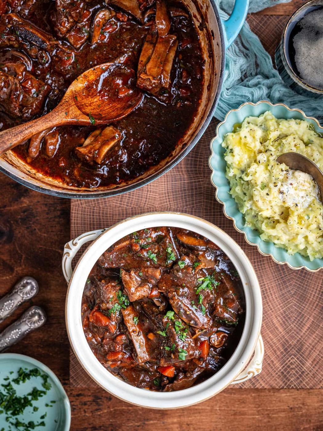 A hearty beef stew with vegetables and tender Guinness short ribs in a white bowl garnished with parsley, beside a pot of stew and creamy mashed potatoes with butter, all set on a wooden table—the ultimate comfort food.