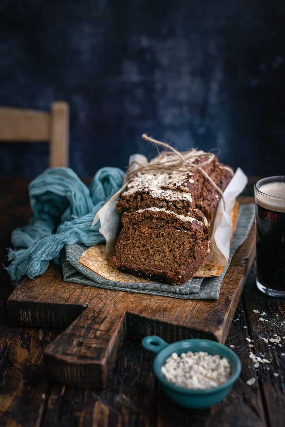 A rustic loaf of Guiness Bread, partially sliced and topped with oats, sits on parchment paper on a wooden board. Nearby are a blue cloth, a small bowl of oats, and a glass of dark beer—perfect for an Irish brown bread recipe​.